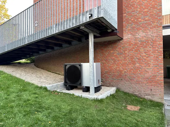 Warmtepomp-buitendeel onder een buitentrap naast een bakstenen woning